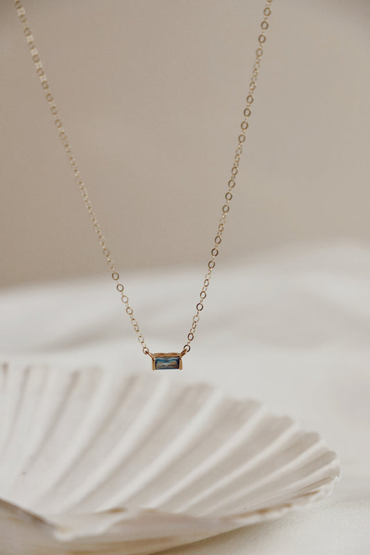 A delicate gold necklace hangs, featuring a rectangular gemstone pendant. It is positioned above a textured, shell-shaped object, set against a soft, neutral background that enhances its elegance.