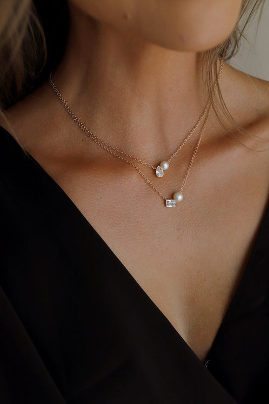 A layered gold necklace adorned with pearls and a small gemstone hangs around the neck of a person wearing a black garment, highlighting the delicate jewelry against their skin.