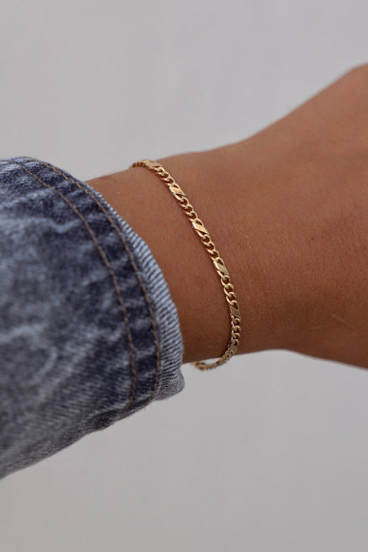 A gold chain bracelet is worn on a wrist, with the hand resting against a neutral background. The bracelet is delicate, featuring alternating links that create a stylish appearance.