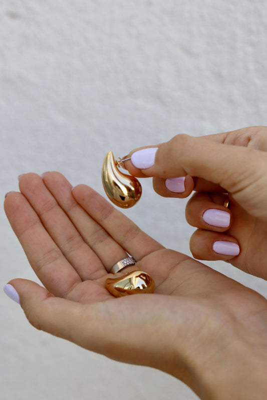 A hand holds a shiny golden earring while another hand cradles a matching piece below. The setting has a simple white background, emphasizing the jewelry’s reflective surface.