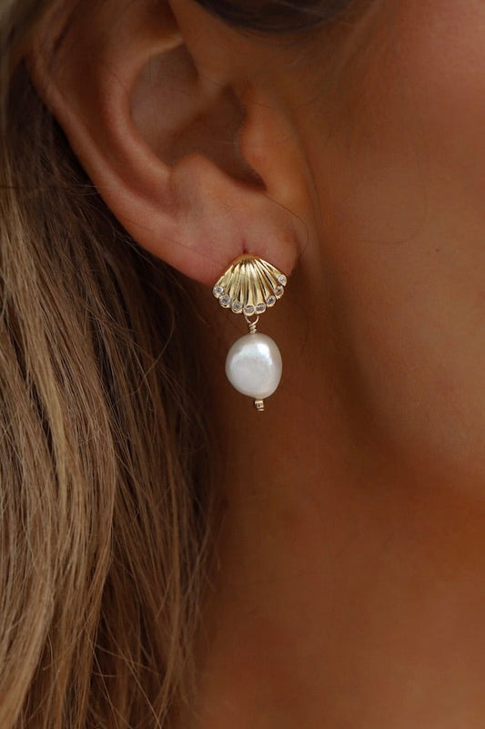 A gold shell-shaped earring features a hanging white pearl, accentuating the ear of a person with long, wavy hair, against a neutral background.