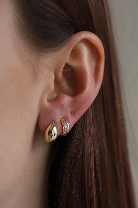 A close-up of a woman's ear showcases two earrings: a shiny gold droplet and a smaller piece with three embedded gems. The ear is partially obscured by long brown hair.