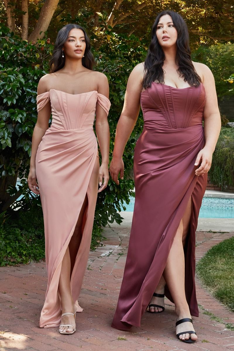 Two women in elegant evening gowns walk together along a brick pathway. One wears a blush dress and the other a deep maroon dress, with a lush garden and pool in the background.