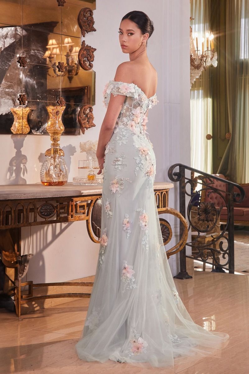 A woman stands gracefully in a light blue, floral-embellished gown, looking back over her shoulder. She is positioned near an ornate table with decorative items in a luxurious, softly lit room.