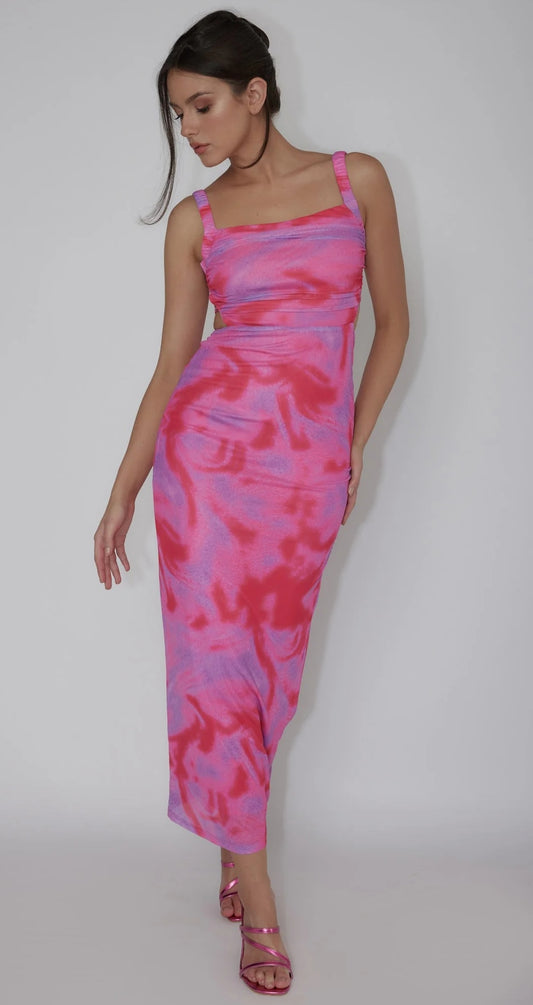 A model poses confidently in a vibrant pink and purple tie-dye maxi dress, one hand on her hip. The neutral background emphasizes her outfit and style.