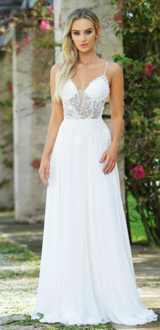 A woman in a flowing white dress stands confidently. The dress features a sheer, embellished bodice and delicate straps, set against a backdrop of greenery and stone walls.