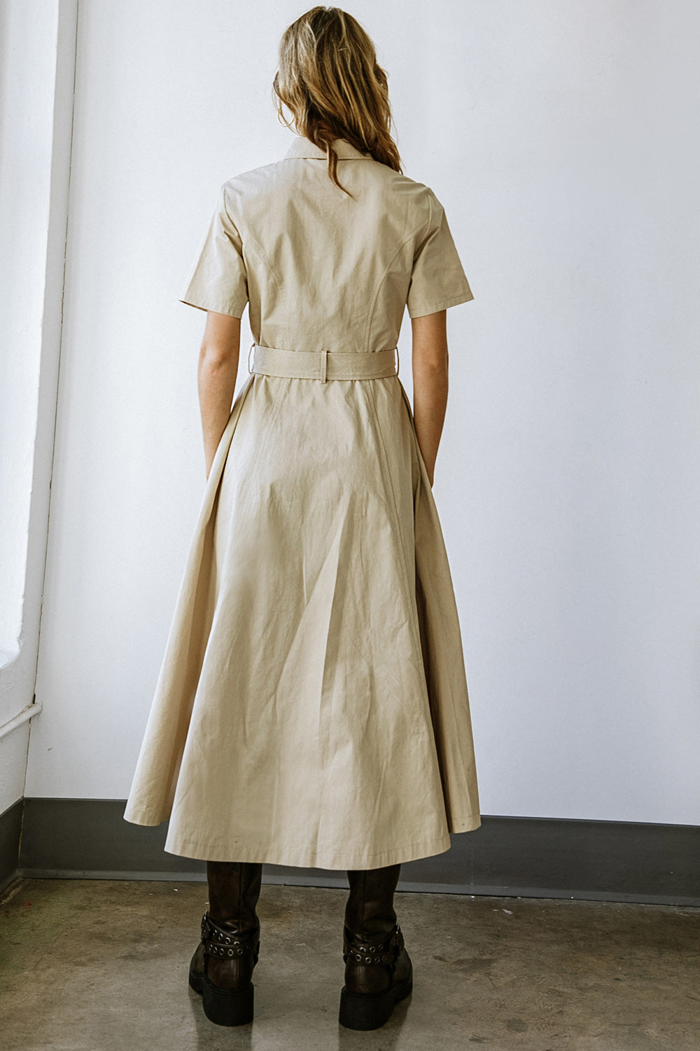 A woman wears a long, beige dress with short sleeves and a belted waist. She stands against a plain white wall in an indoor space with a concrete floor.