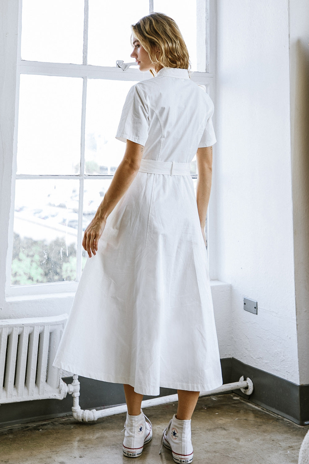 A woman wears a flowing white dress with short sleeves and a belt, standing by a large window. She faces away, revealing a casual style complemented by sneakers, in a well-lit room.