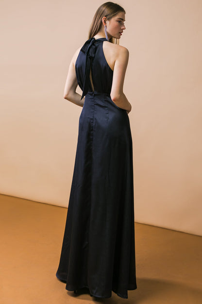 A model stands in a black, sleeveless gown with a halter neck and a flowing skirt. The backdrop is a soft beige, while the floor is a warm brown.