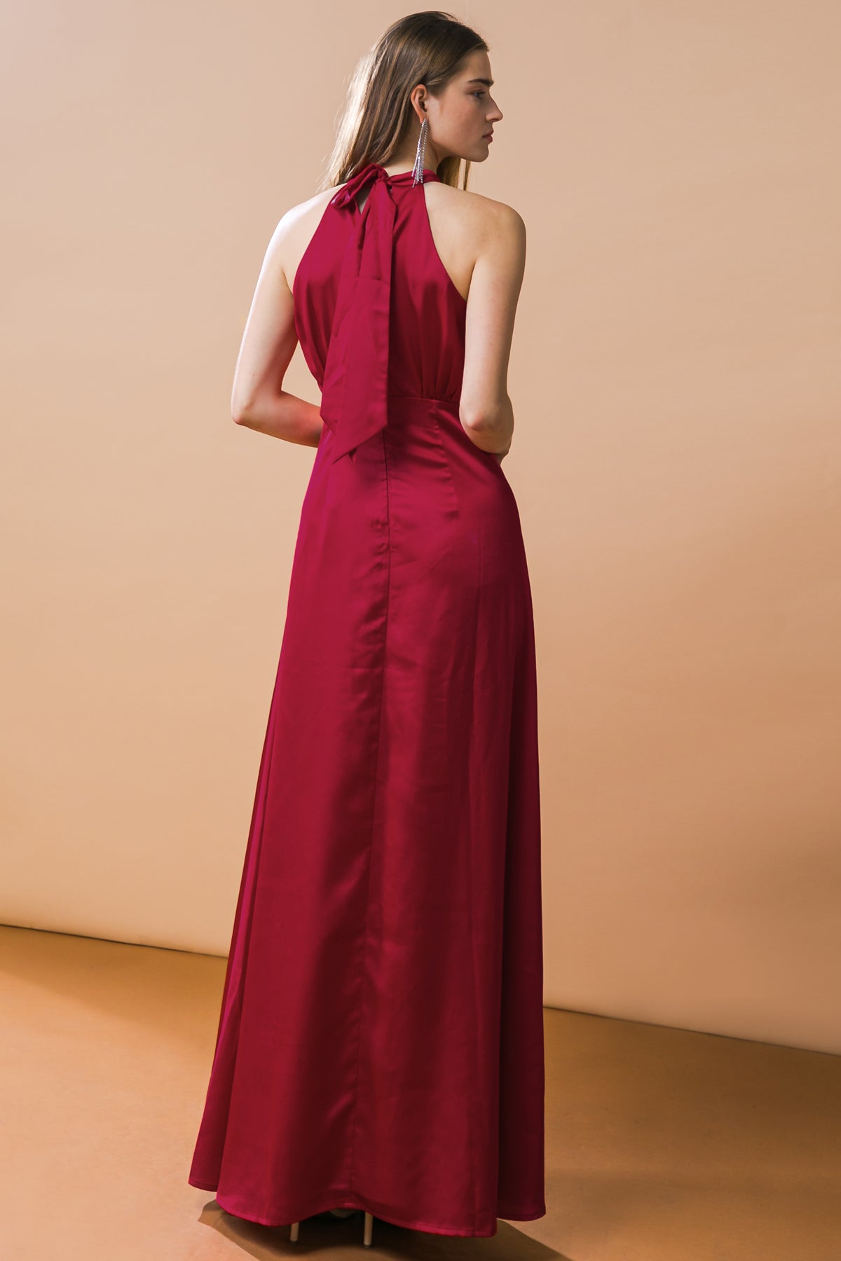 A woman models a sleeveless red gown with a tied neck, standing against a smooth, neutral-colored backdrop. The dress flows down to the floor, creating an elegant silhouette.