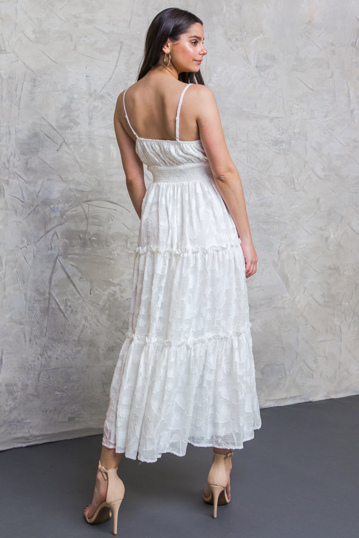 A woman stands gracefully, wearing a flowing white dress with a tiered design. She poses with her back facing the camera, against a textured, light-colored background, showcasing her stylish heels.