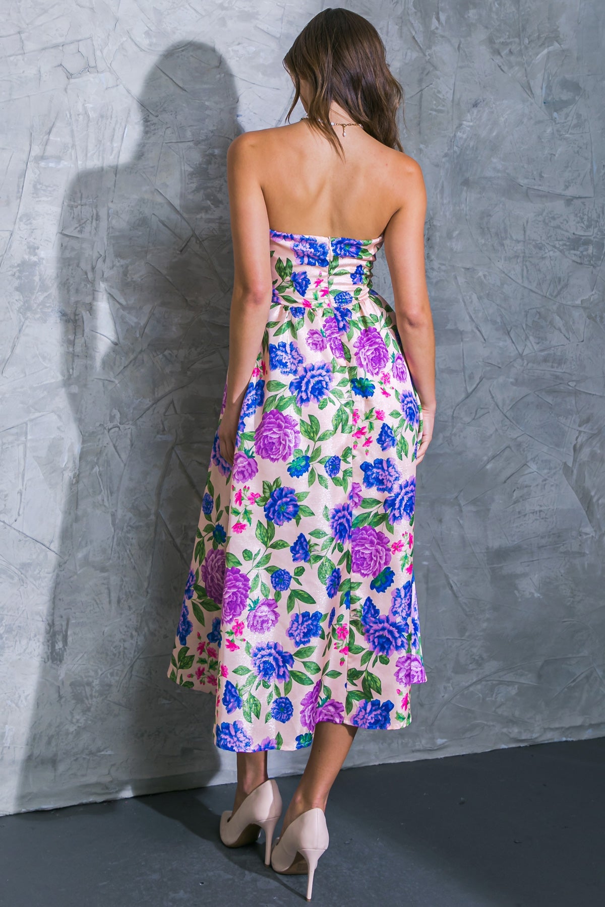 A woman stands with her back to the viewer, wearing a strapless floral dress with large purple and blue flowers. She is in front of a textured gray wall, and her shadow is visible.