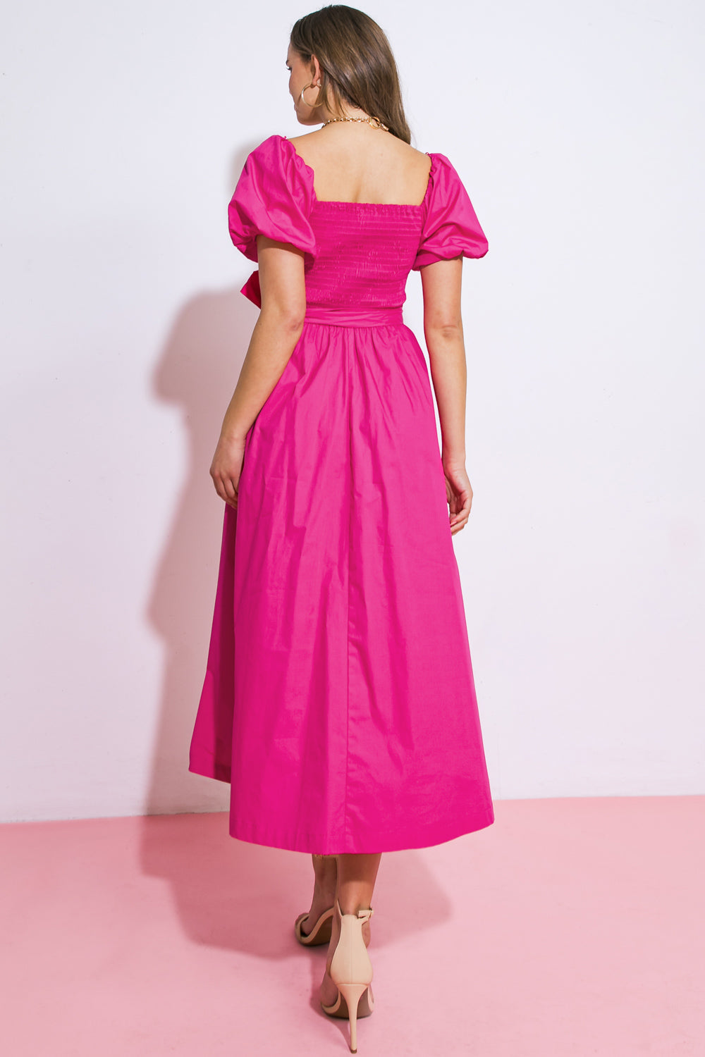 A person wearing a bright pink, puff-sleeved dress stands with their back to the viewer on a pink floor, set against a plain white wall, showcasing the dress's flowing silhouette.