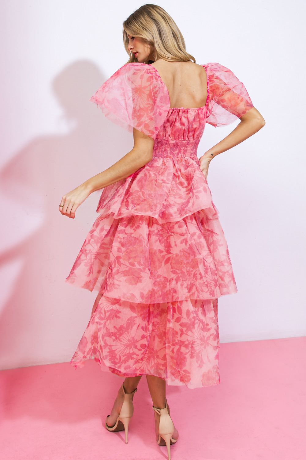 A woman wearing a tiered, pink floral dress with puff sleeves turns gracefully. The background features a soft pink wall and floor, enhancing the dress's vibrant colors.