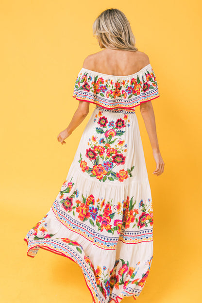 A woman in a colorful, floral-embroidered white dress strolls gracefully, showcasing vibrant patterns against a solid yellow background, enhancing the garment's liveliness and style.