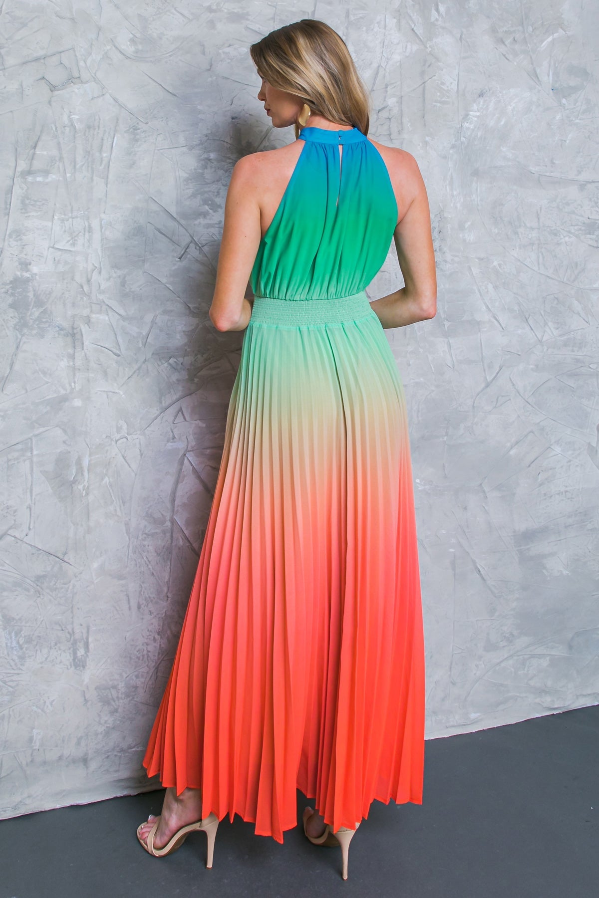 A woman wears a vibrant, pleated maxi dress with a gradient from green to orange, standing against a textured gray wall. Her stylish sandals complement the outfit.