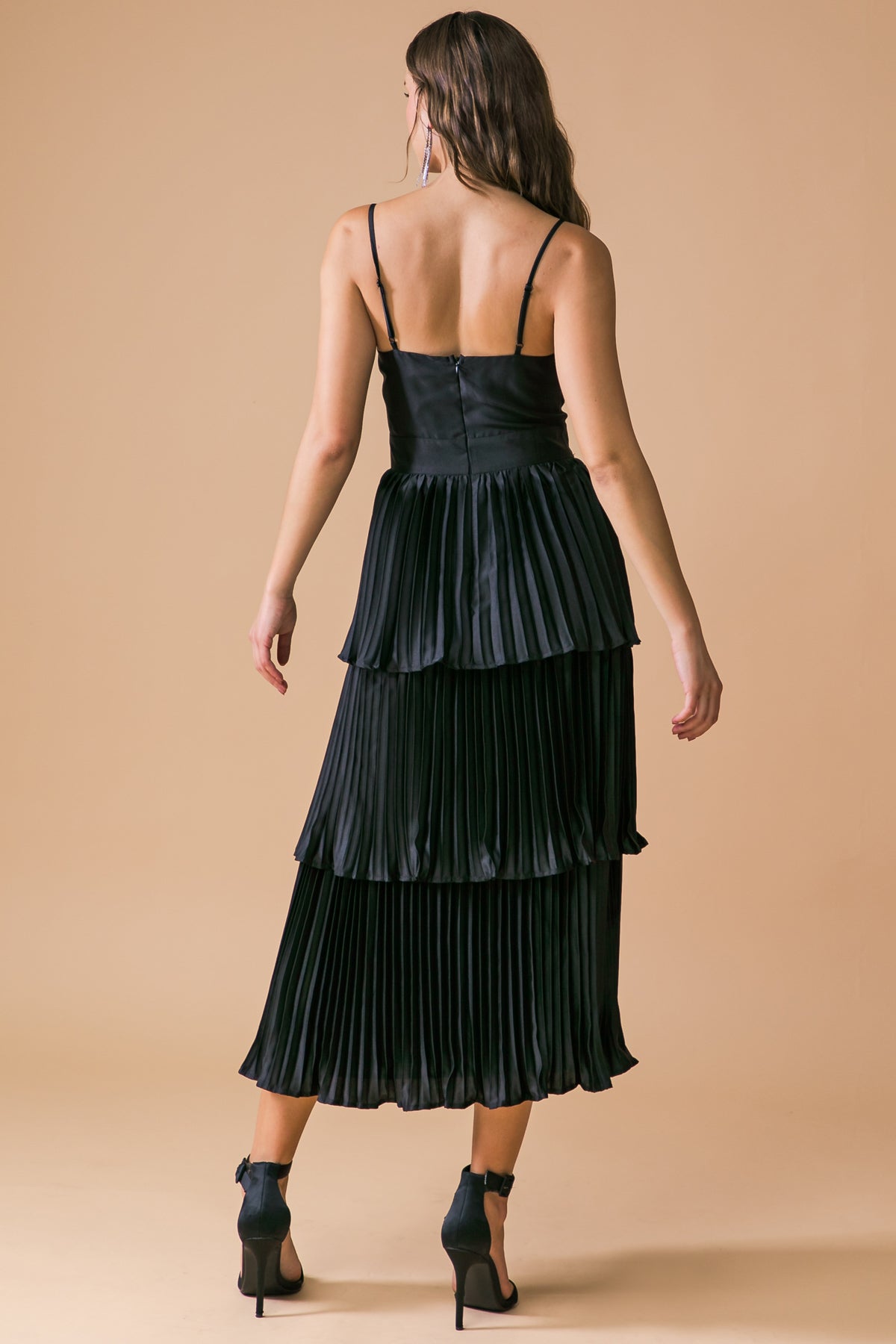 A black, pleated midi dress with thin straps is worn by a model, who stands in profile against a smooth, beige background, showcasing the dress's layered design and elegant silhouette.