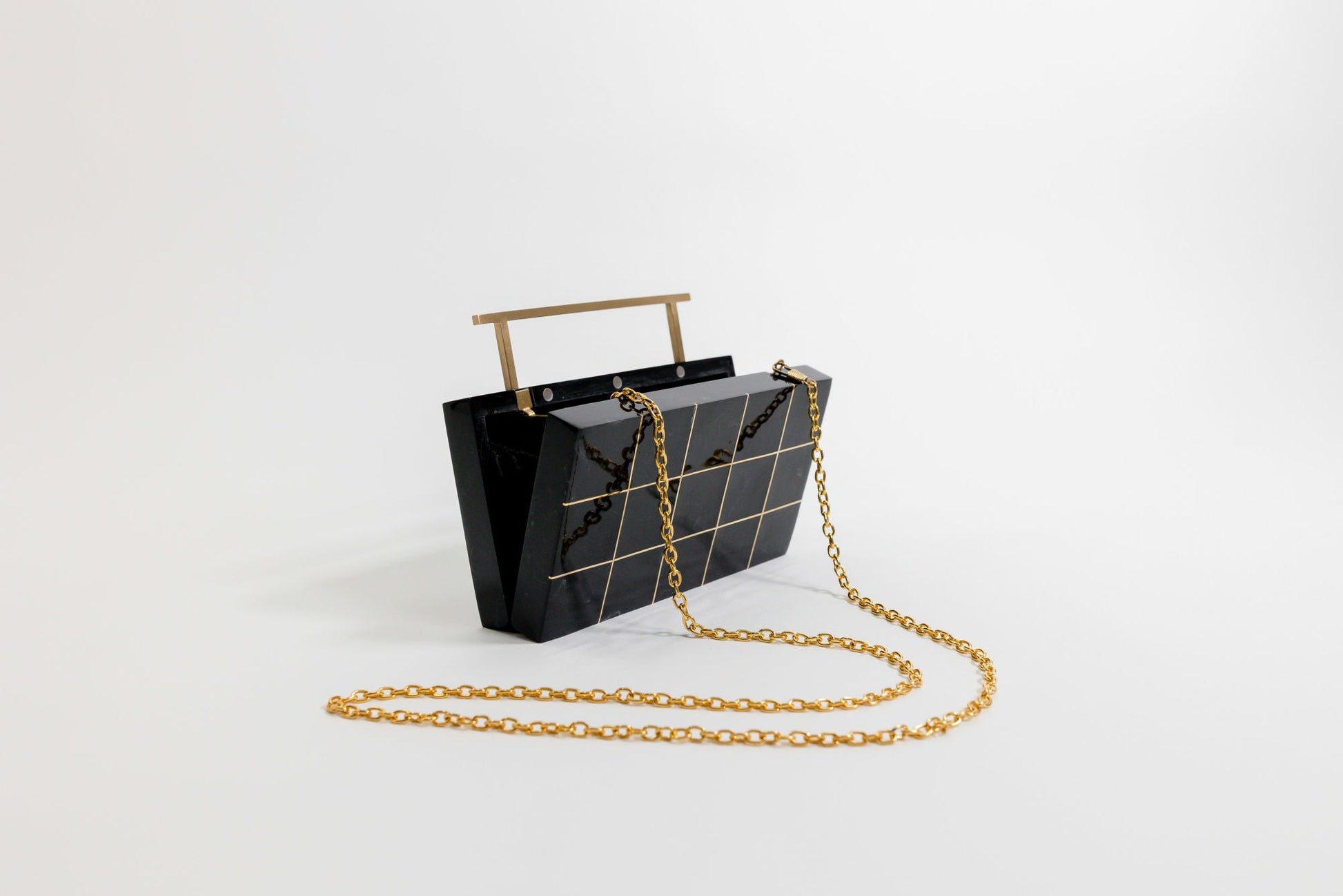 A black, rectangular handbag with gold chain strap features grid-like patterns. It stands upright on a plain white surface, with a horizontal handle at the top.