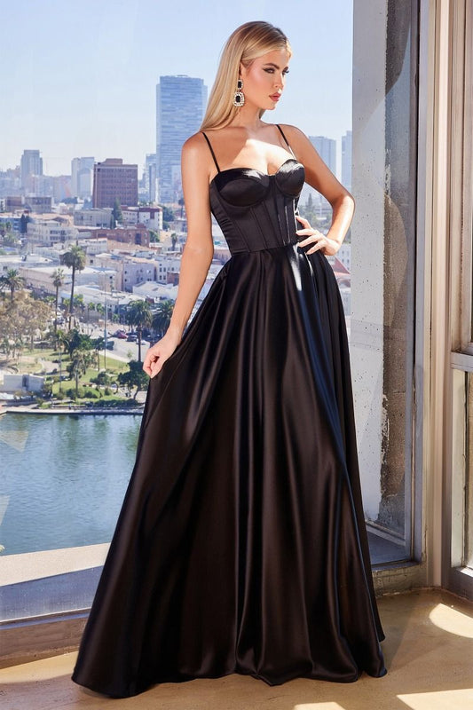 A woman stands elegantly in a black, floor-length gown with a fitted bodice and thin straps. She poses confidently by a large window, showcasing a city skyline and water view behind her.