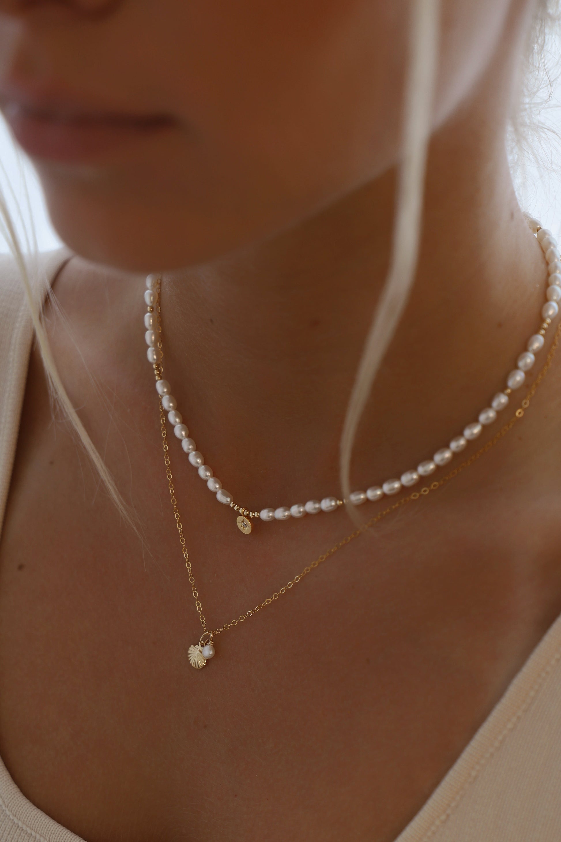 A woman displays two delicate necklaces around her neck. The top necklace features pearls, while the lower one has a charm with a small pearl, set against a softly lit background.