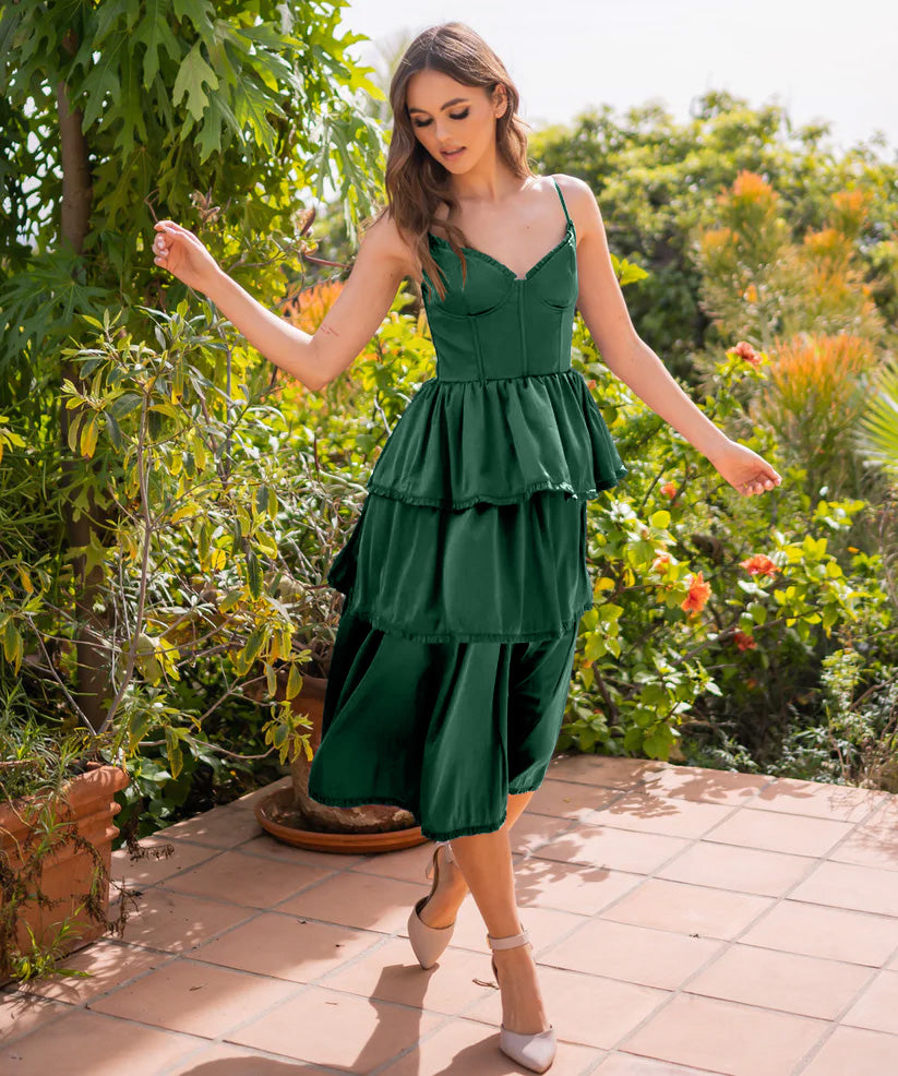 A woman in a green layered dress gracefully walks on a tiled patio. Surrounding her is lush greenery and colorful flowers, creating a vibrant and sunny outdoor setting.