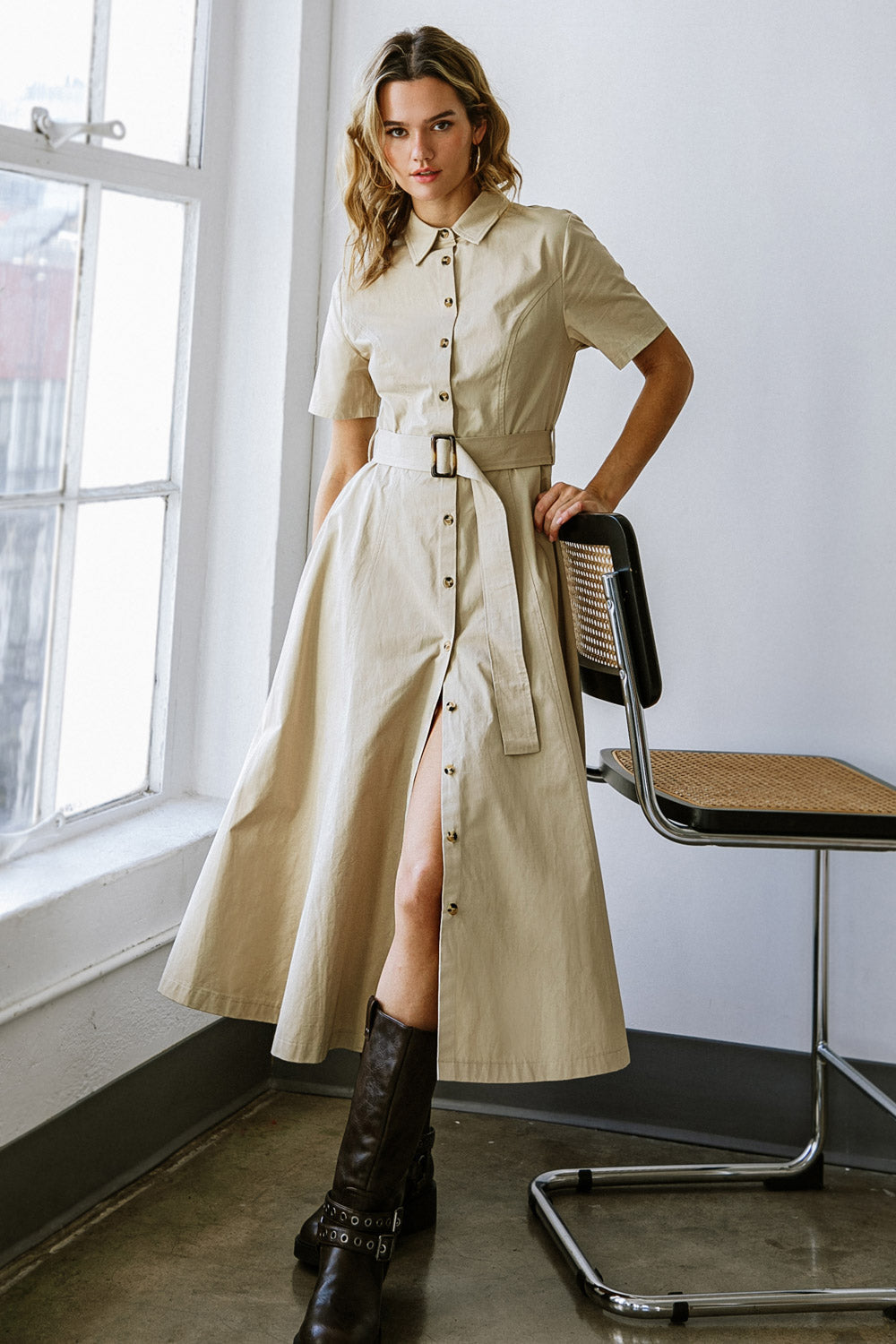 A woman wears a khaki shirt dress with a belt, standing confidently beside a modern chair. Sunlight streams through a large window, illuminating the minimalist indoor space.