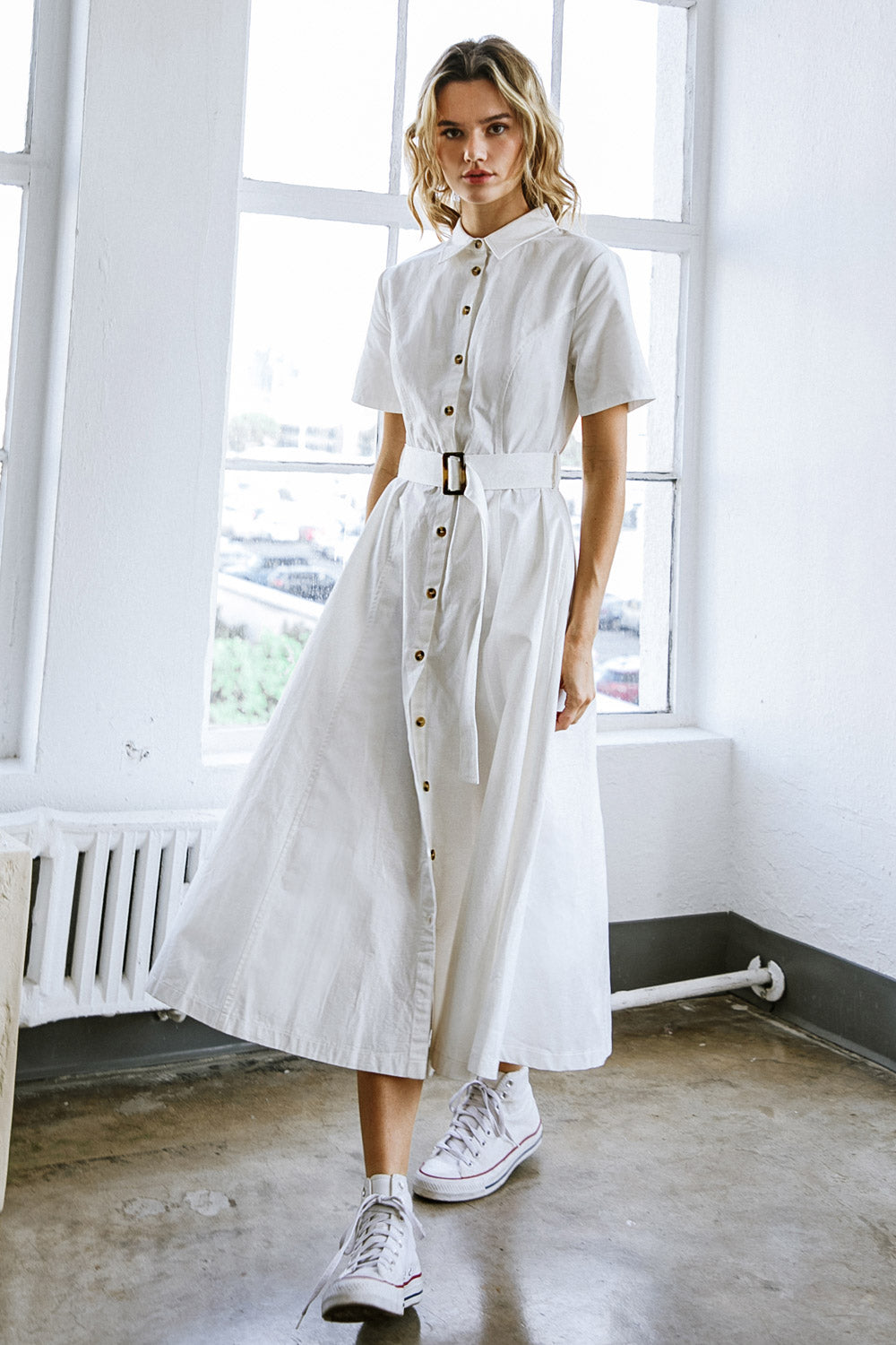 A model stands in a bright, minimalist room wearing a long, white button-up dress with a belt, paired with white sneakers. Sunlight illuminates the space through large windows.