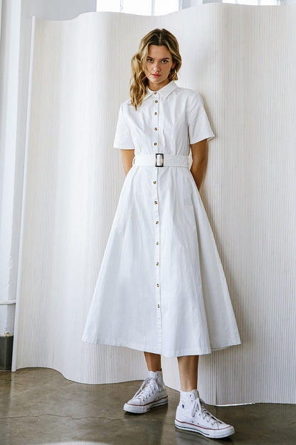 A woman wears a white, button-down dress with a belt, standing against a textured backdrop. She pairs the dress with white high-top sneakers, showcasing a casual yet stylish look.