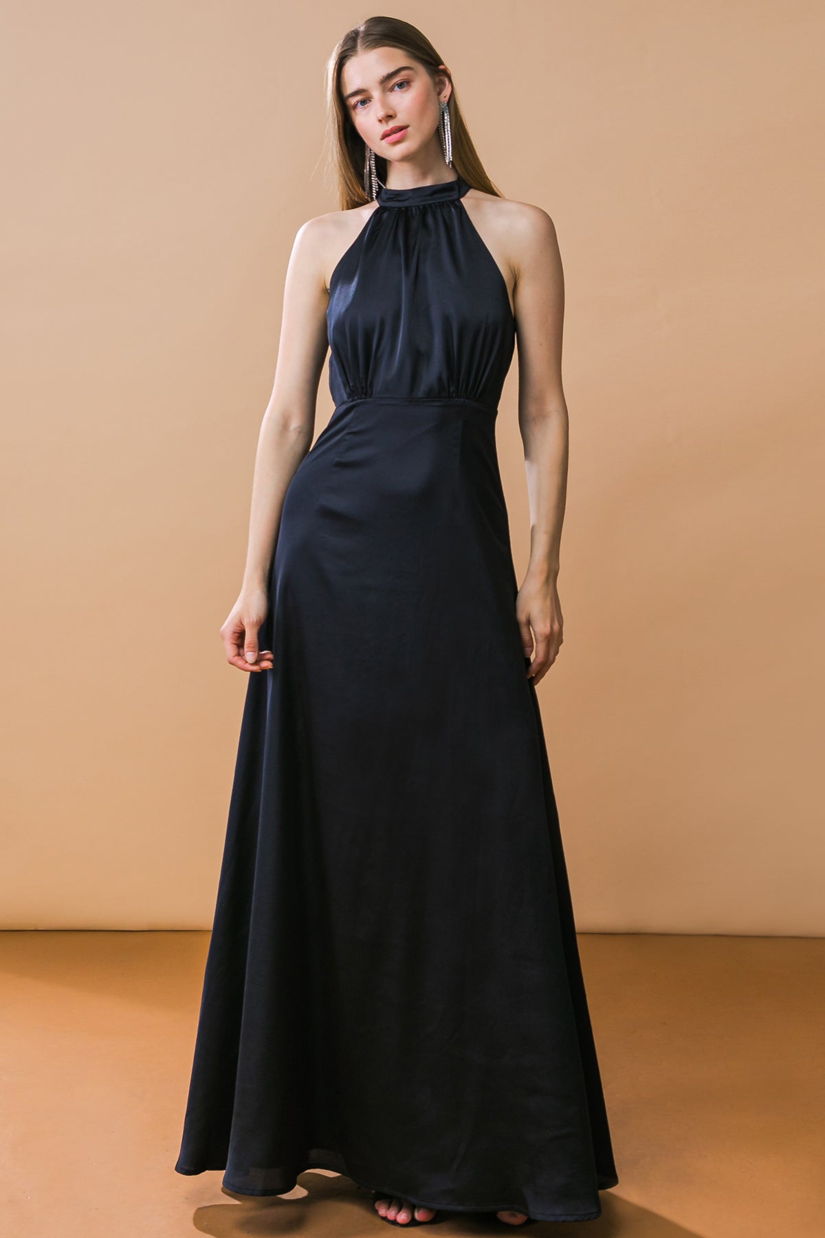 A model stands confidently in a sleek black halter-neck gown that flows to the floor. She poses against a warm, orange backdrop, accentuating her elegant look.