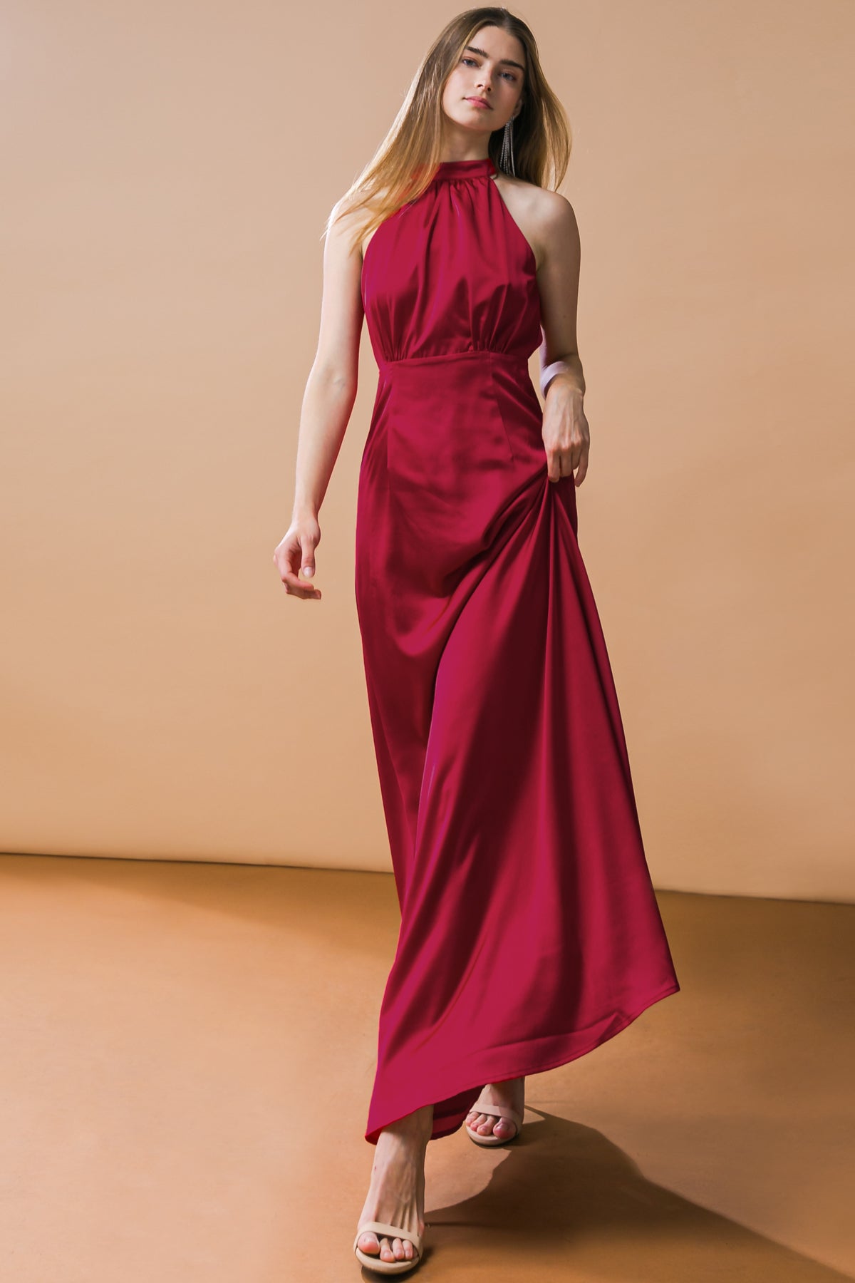 A woman in a flowing red halter dress walks confidently forward, her long hair cascading over her shoulders, against a soft beige background and a warm brown floor.