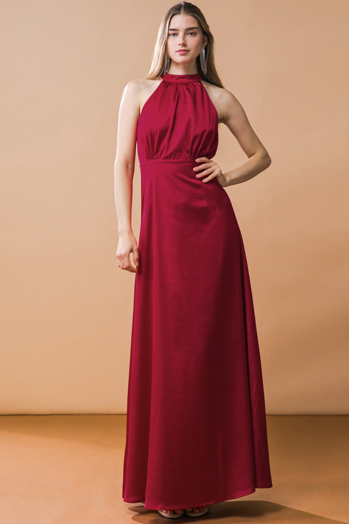 A woman stands confidently in a flowing, elegant burgundy halter dress on a smooth, light-colored floor. The backdrop features a warm tone, enhancing the dress's rich hue.