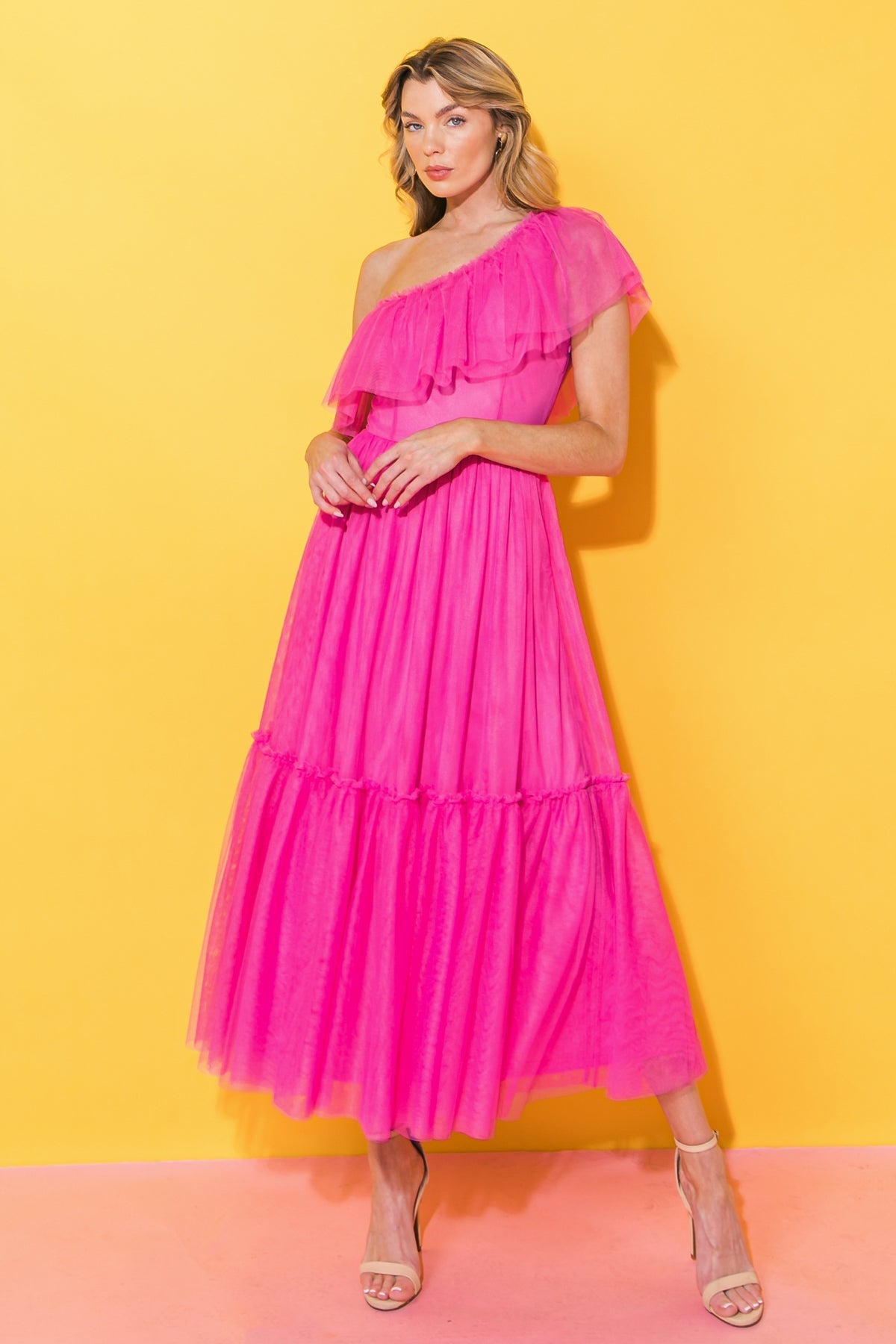 A woman poses confidently in a vibrant pink dress with ruffles, standing against a bright yellow background. She wears stylish heels, enhancing the bold fashion statement.