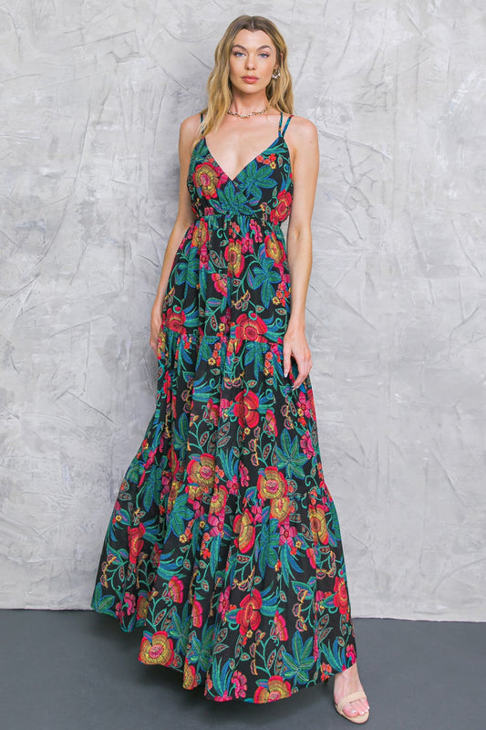 A woman stands confidently, wearing a vibrant, floral-patterned maxi dress. The dress features colorful flowers on a black background, complementing her relaxed pose against a textured gray wall.