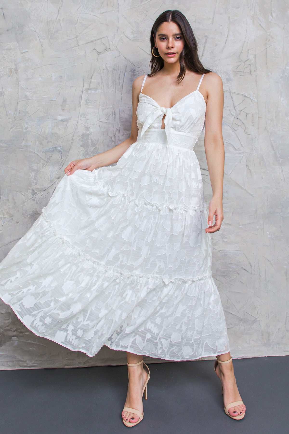 A woman is playfully lifting the hem of her white, tiered maxi dress, showcasing its floral texture. She stands against a neutral, textured wall, wearing beige strappy heels.