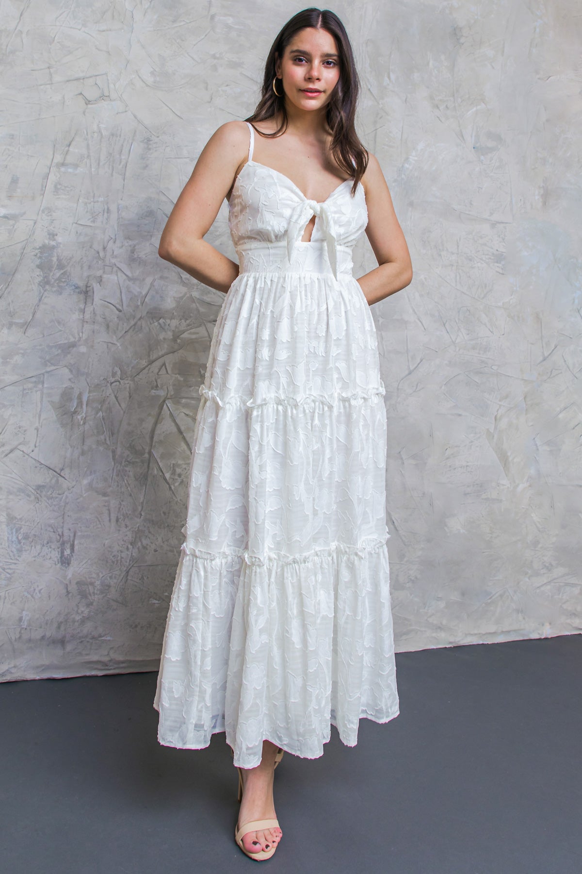 A woman stands confidently, wearing a flowing white dress with a tie front and layered ruffles. The backdrop features a textured gray wall, creating a soft, elegant atmosphere.