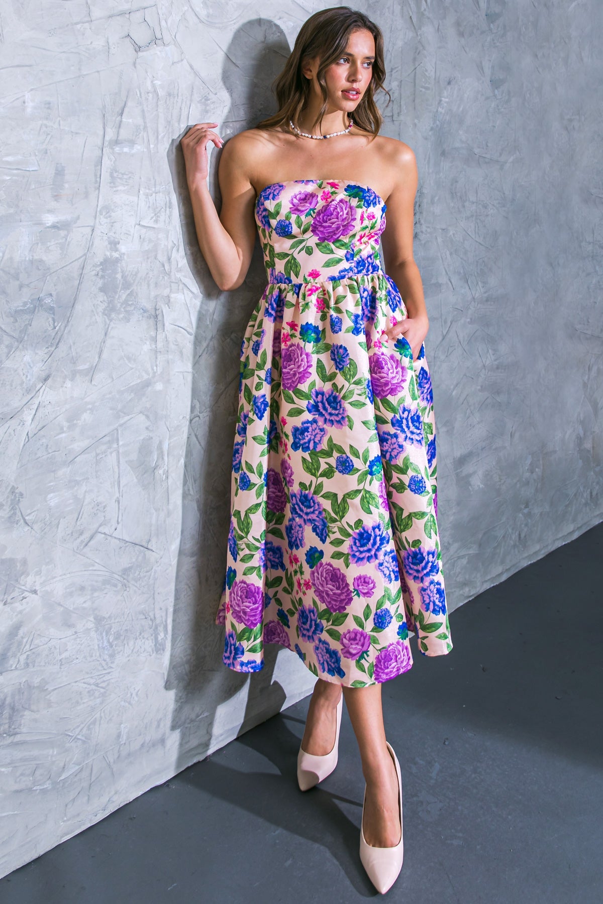 A woman in a strapless floral dress leans against a textured gray wall, exuding elegance and style, showcasing vibrant purple and blue flowers against a light background. She wears pink high heels.