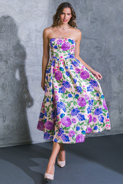 A young woman wears a colorful floral dress, smiling as she walks gracefully. The setting features a textured gray wall, enhancing the vibrant colors of the dress and the soft lighting.