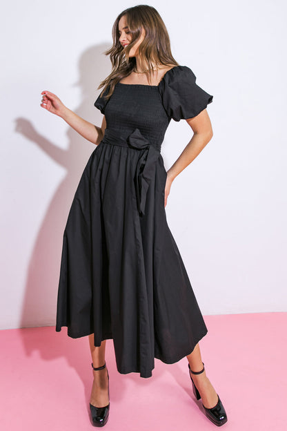 A woman in a black, long, puff-sleeved dress stands gracefully, with one hand raised. The background features a white wall, and the floor is a soft pink hue, creating a stylish contrast.