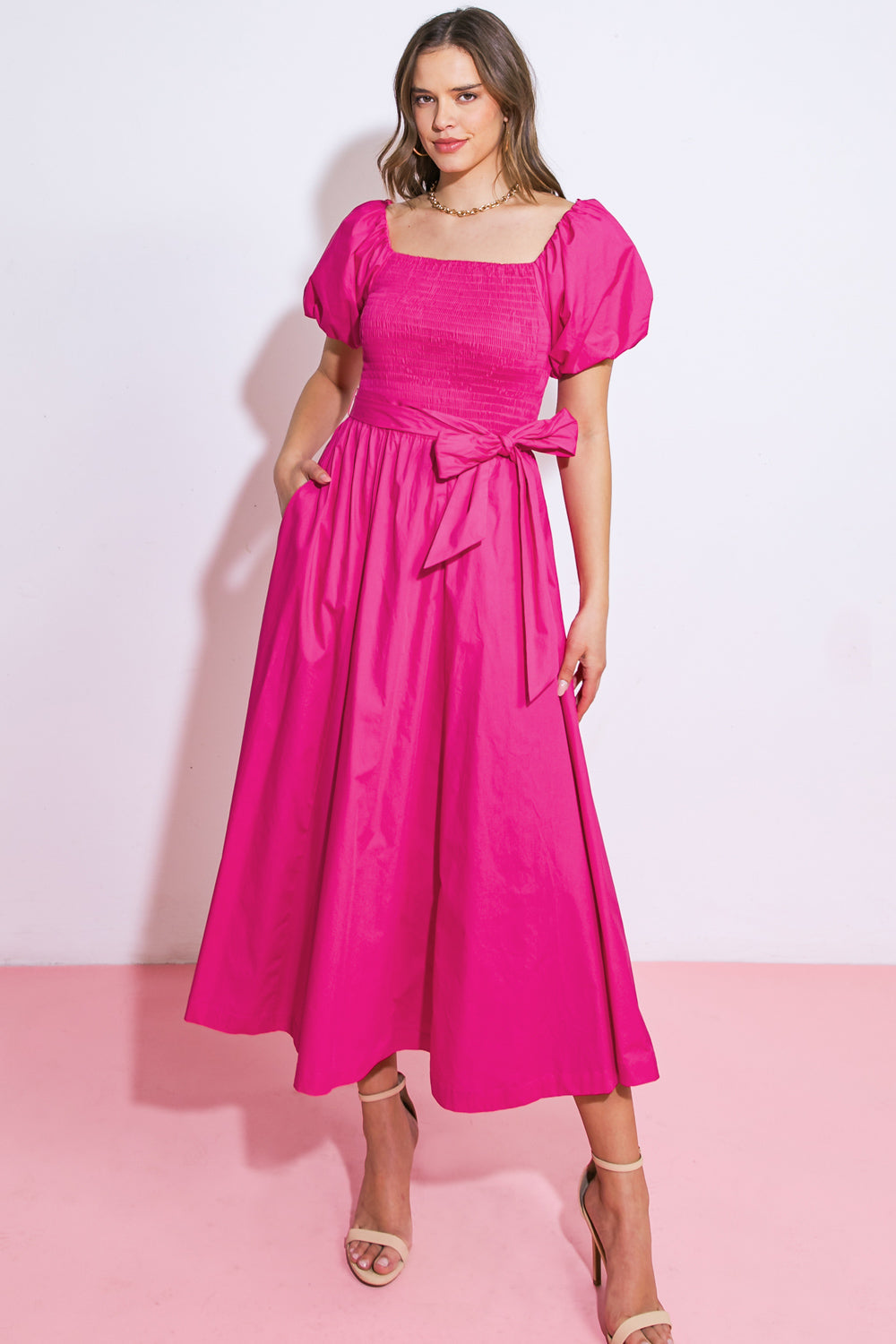 A woman stands confidently, wearing a vibrant pink dress with puffed sleeves and a tied waist. She poses against a soft pink background, showcasing a stylish and playful look.