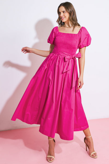 A woman wearing a vibrant pink dress with puff sleeves stands gracefully, holding her arm out. The setting features a light pink floor and a plain white wall, enhancing the dress's color.