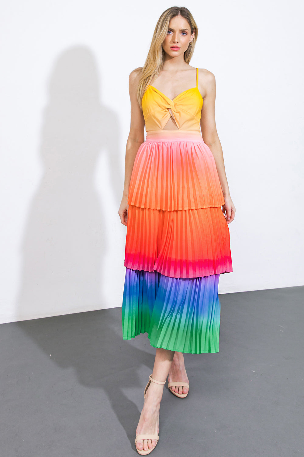 A woman stands confidently wearing a colorful, pleated dress with a yellow top and a tiered skirt blending hues from orange to blue. The background is plain white, and the floor is grey.