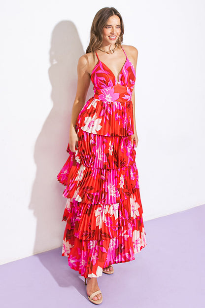 A woman stands smiling, wearing a vibrant, sleeveless red dress with pink and white floral patterns and layered ruffles, against a white wall and purple floor.