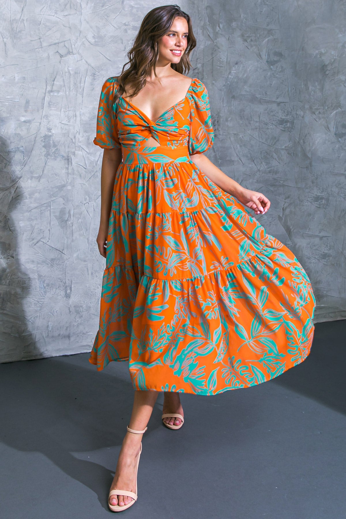 A woman in an orange, floral-patterned dress twirls gracefully, showcasing the dress's flow and design. She stands on a dark floor against a textured gray wall backdrop.