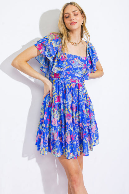 A woman wearing a blue floral dress poses with one hand on her hip and a slight smile, set against a plain white background. Her hair is loose, and she accessorizes with gold jewelry.