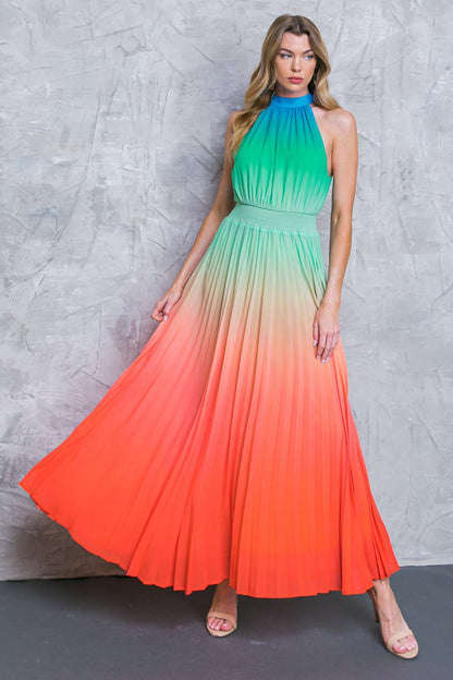 A woman stands elegantly in a long, pleated dress that transitions from green at the top to orange at the bottom. The background features a textured gray wall.