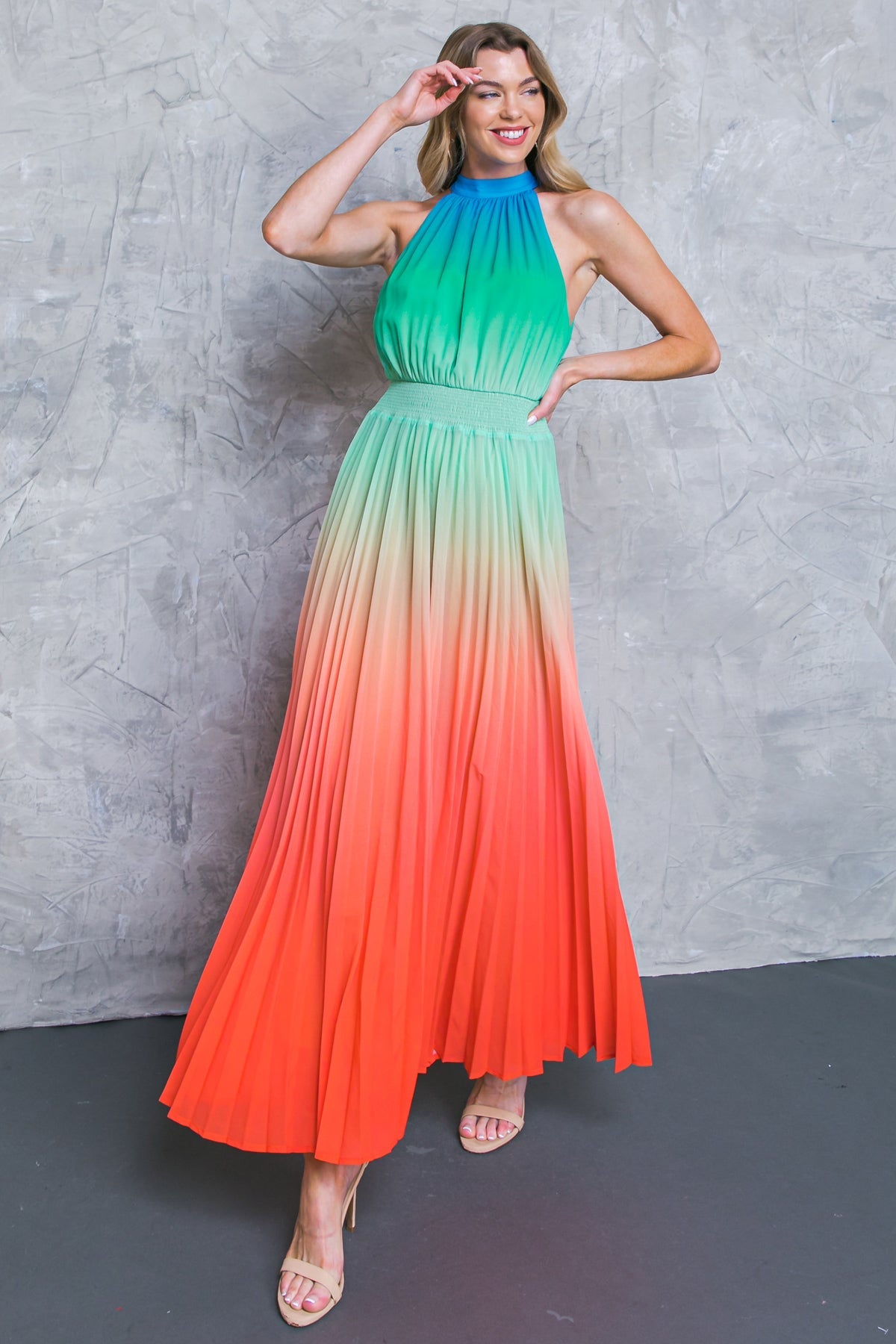 A woman in a vibrant halter dress transitions from green to orange, striking a pose with one hand on her head, against a textured gray wall. She wears beige high heels.