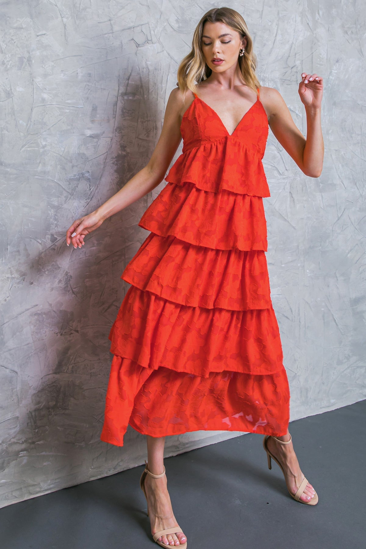 A woman in a bright red, tiered dress poses gracefully against a textured gray wall, one hand raised slightly, showcasing the dress's playful layers and her strappy beige heels.