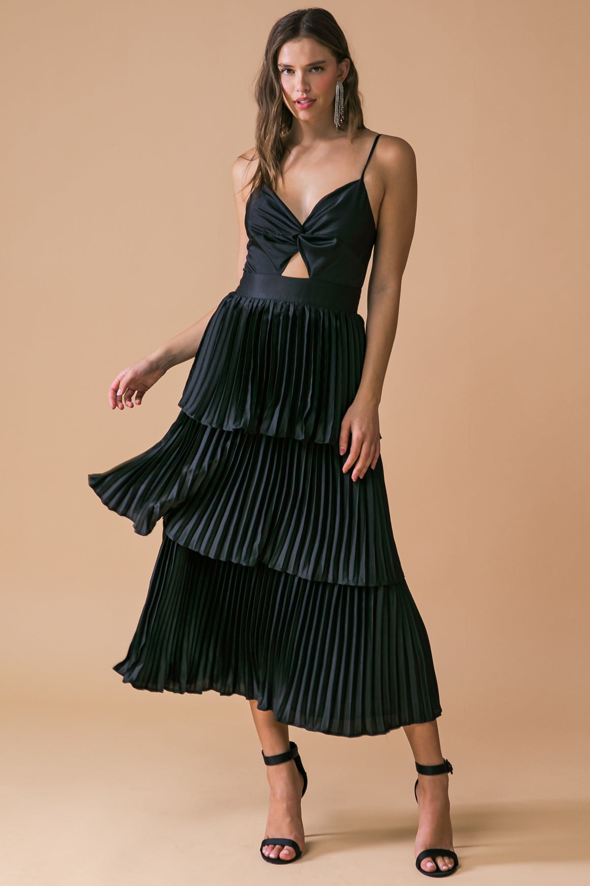 A woman wearing a black pleated dress with a twisted bodice and a cutout stands gracefully, one hand lifted slightly as the dress flows around her, against a soft beige background.