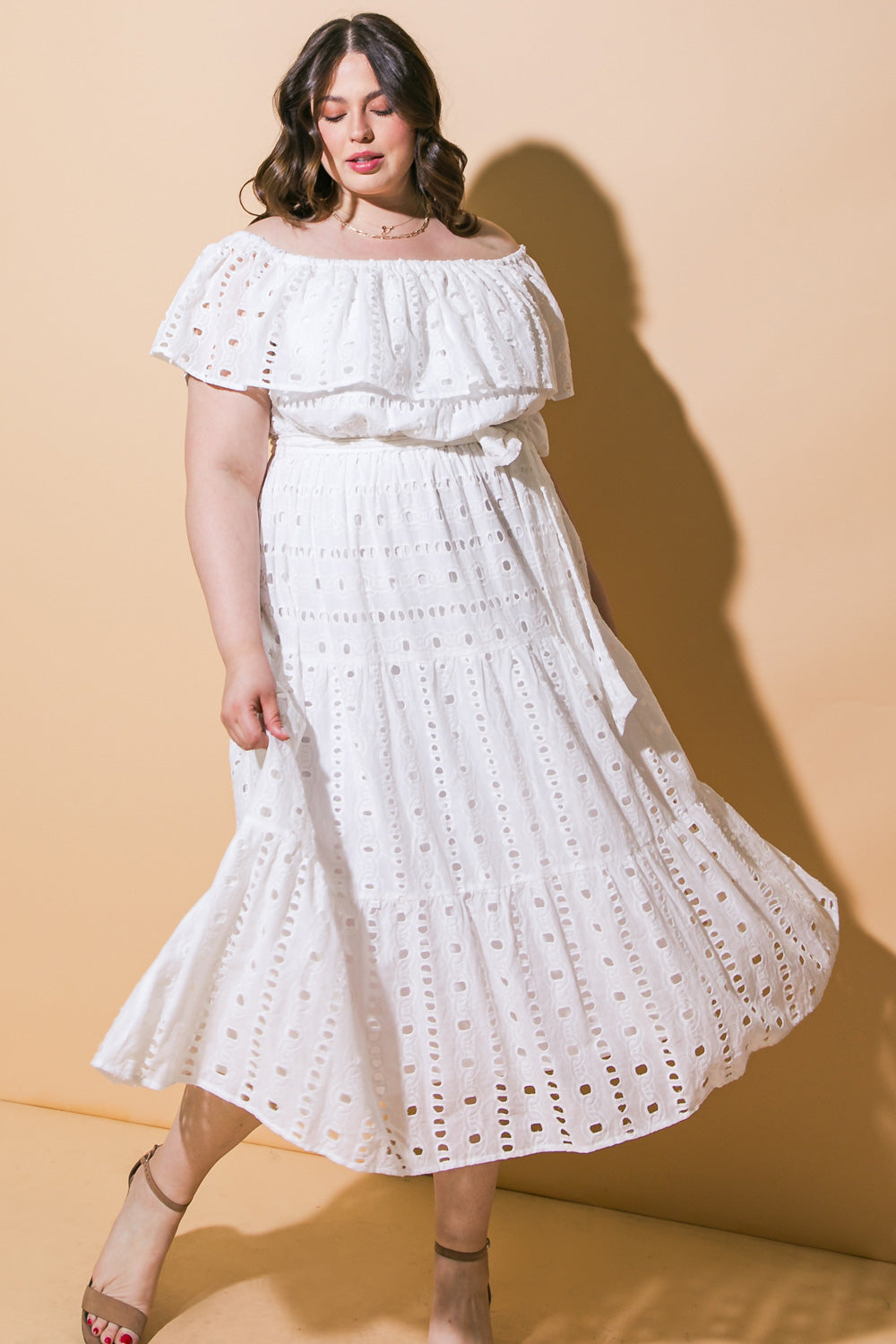 A person wearing a white off-shoulder dress with eyelet patterns stands gracefully, holding part of the dress, against a soft yellow backdrop, creating a warm and inviting atmosphere.