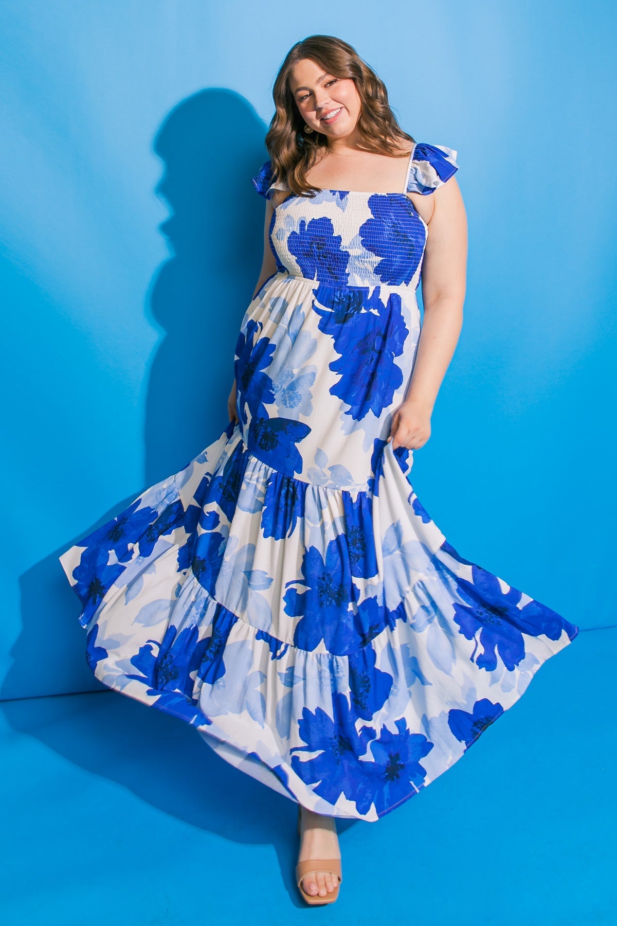 A woman stands smiling, wearing a flowing, blue floral dress, twirling slightly. She is in front of a bright blue background, which enhances the vivid colors of her outfit.